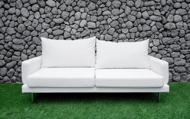 A white couch is sitting on a green lawn in front of a stone wall. The couch is empty and the wall is made of rocks