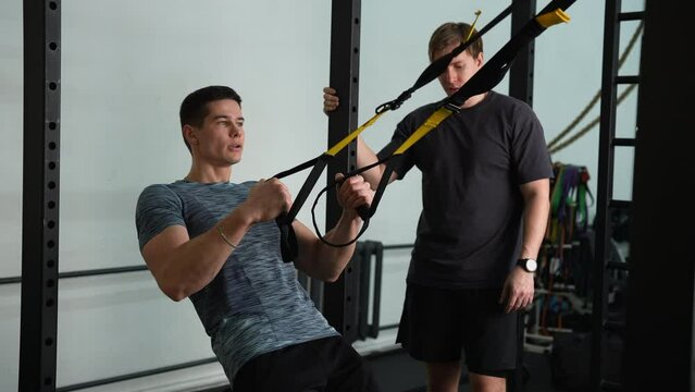 Muscular fitness instructor male showing suspension training to motivated beginner young man, performing pull-ups exercise with trx straps at gym. Lifting up and down to maintaining muscles.