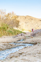 A small stream and rocky mountains with tree, bushes and red colored granite rocks. An unrecognizable person walking to a 4x4 car. Surface level view, reflective flowing water in an arid desert land.