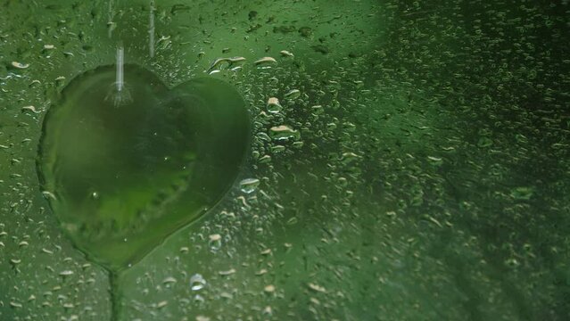 Window car pane with rain drops falling down, making heart shape, against background of green nature.