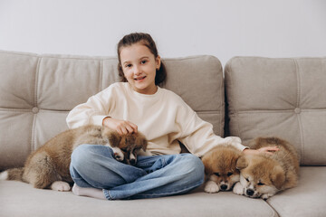 Little happy smiling girl playing and hugging Akita Inu puppies at home on sofa