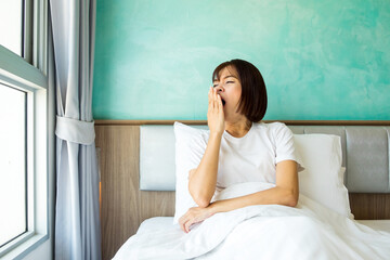 A young Asian woman in a white t-shirt sits and yawns with her hand over her mouth on a bed with...