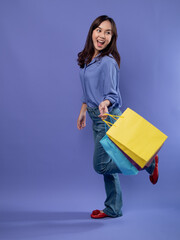 Joyful shopper with colorful bags, blue blouse, denim jeans, and red flats on a purple backdrop