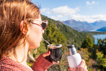 Woman enjoys some delicious mate with yerba while observing the most beautiful landscapes of southern Argentina, surrounded by mountains and lakes. Bariloche, Patagonia, Argentina.