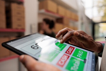 Worker checking online customers orders using tablet computer, preparing cardboard boxes before...