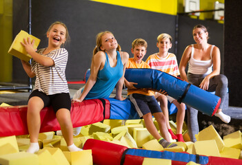 Group of happy tween children and young woman sitting on soft beam above pit of foam cubes,...