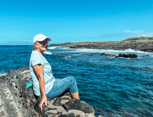 Senior woman in outdoors vacation at sea sitting on a rocky beach admiring waves splashing on the...
