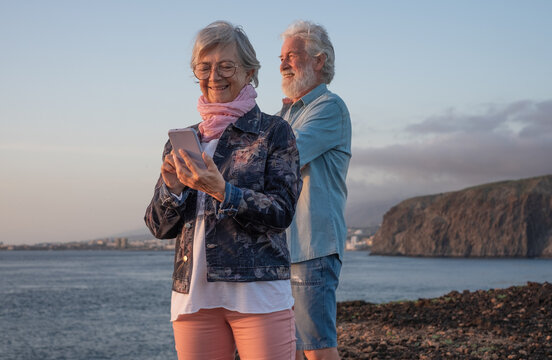 Smiling senior couple standing on the rocky beach at sea enjoying vacation and retirement, man looking at horizon over water, woman using phone