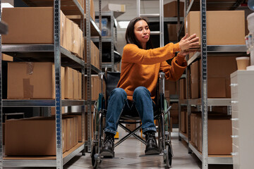 Woman wheelchair user taking cardboard box from shelf to prepare customer order for dispatching in...