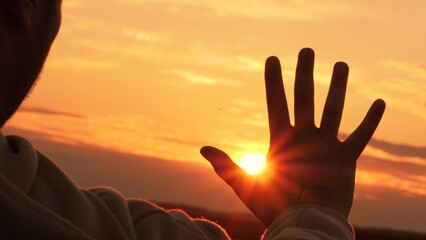 Silhouette of hand at orange sunset. Man stretches hand towards sun, warming palm in summer rays....
