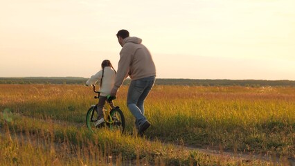 Parent teaches child to ride bicycle. Father and daughter in field at epic sunset. Child safety,...