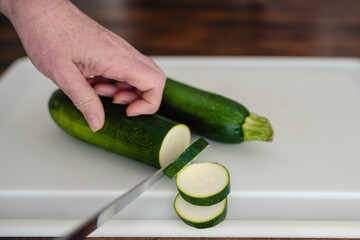Close-up of older woman's hands slicing zucchini with a knife on a white cutting board horizontal