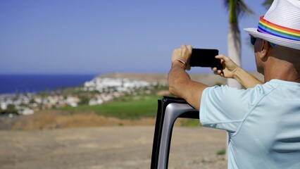 Young gay hispanic man with rainbow bracelet, hat and sunglasses, photographs the resort outside...