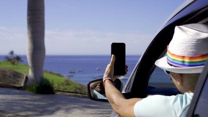 Young gay tourist wearing rainbow bracelet and hat leans out of car window to photograph coastal...