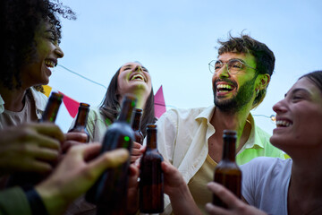 Portrait of diverse young friends laughing toasting with beer bottles celebrating party on a summer evening outdoor. Excited happy millennial people gathering enjoying music at rooftop terrace