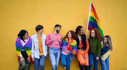 Group of diverse LGBT young people standing on wall yellow. Smiling friends celebrating gay pride day enjoying together and showing rainbow flags. Generation z relationships and open mind