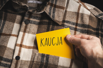 Yellow card with a handwritten inscription "Kaucja", held in the hand against the background of a brown plaid shirt (selective focus), translation: deposit