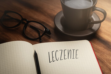 A handwritten inscription "Leczenie" on a grille of an open notebook on a wooden countertop, next to a black pencil, a cup with coffee and glasses, a flash of light. (selective focus), translation: tr