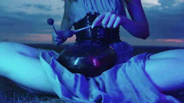Young performer plays the hapi drum in blue neon light, against the backdrop of an inspiring and meditative evening sky. She are sitting on the grass, in the lotus position, surrounded by nature