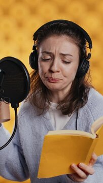 Vertical video Voice actor reads book while enjoying coffee, recording audiobook using microphone, studio background. Woman using mic and headphones to produce digital recording of novel, drinking