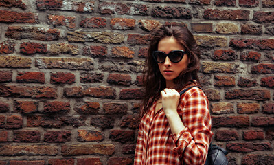 Beautiful brunette woman with long brown hair in fashion sunglasses standing on the old red brick wall building background in casual red shirt on the spring city. Closeup vintage banner - 794518822