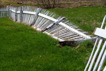 Closeup of broken white picket fence with rotted wood post lays fallen in green grass