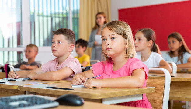 Diligent elementary school student preteen girl studying with classmates, making notes of teacher lecture