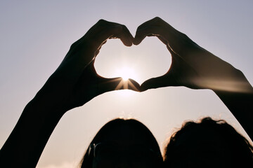 Silhouette, heart hands and couple at sky for sunset, love and care together on summer holiday with...