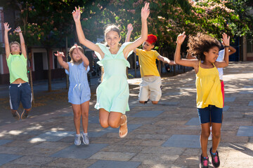Obraz premium Happy tween girl having fun outdoors with friends of different nationalities, jumping on city street in summer day..