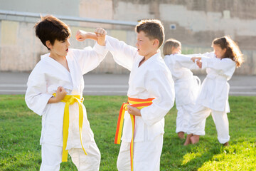 Focused school children wearing white sports uniform practicing karate sparring in pairs on green...