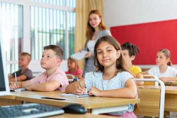 Portrait of smiling tween schoolgirl sitting in classroom during lesson, writing exercises in workbook