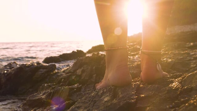 LENS FLARE, CLOSE UP: Young barefoot lady stands on a seashore where the golden rays of setting sun reflect off the rough wet rocks. Her ankles are adorned with simple and beautiful shell bracelets.