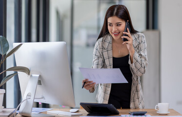 Asian woman entrepreneur busy with her work in the office. Young Asian woman talking over phone or cellphone while sitting at her desk.
