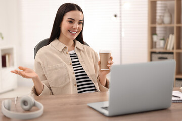 Young woman with cup of coffee using video chat during webinar at table in room