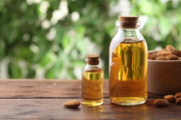Almond oil in bottles and nuts on wooden table against blurred green background, closeup. Space for text