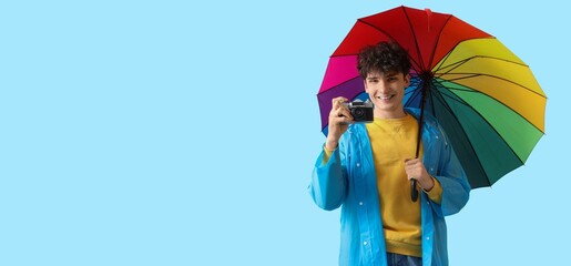 Young man in raincoat holding rainbow umbrella and photo camera on light blue background with space...