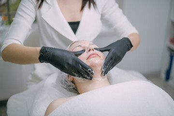Cosmetologist and dermatologist cleans the face of a lying woman patient in a beauty salon. Young...
