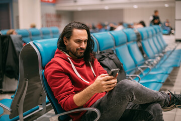 A young, handsome man is sitting with a phone in his hands at the airport in the evening. A guy, a...