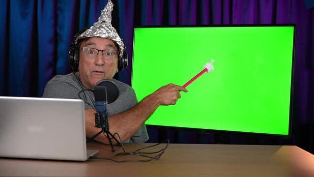A crazy UFO conspiracy theorist with a tin foil hat records a podcast in his home studio. Video monitor to his side with a green screen for your custom content.  	