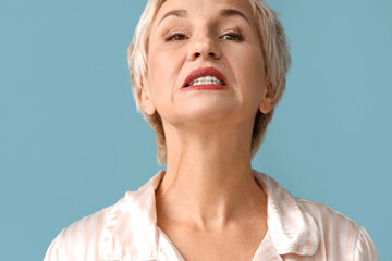 Beautiful mature woman in pajamas doing face building exercise on blue background, closeup