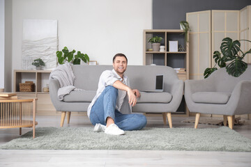 Young man sitting beside grey sofa with laptop at home