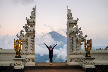 A girl traveler stands at a traditional Balinese gate and admires the view of Agung volcano, rear...