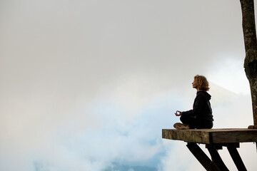 A young girl of European appearance meditates and does yoga on a wooden platform in the mountains,...