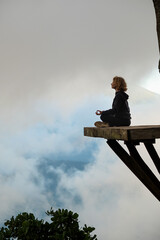 A young girl of European appearance meditates and does yoga on a wooden platform in the mountains,...
