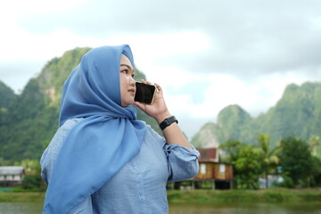 An Asian model in a hijab wearing a blue shirt is posing in nature with a backdrop of mountains and water in the countryside while holding a mobile phone