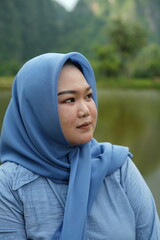 Asian model in hijab wearing blue shirt is posing in nature with mountains and water in the countryside as a backdrop