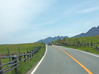 Travelling on rural Kyushu, driving to distant Mount Aso famous volcano caldera contour on lonely road with lush green grass field and wooden fence on roadside