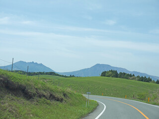 Travelling on Kyushu, driving to distant Mount Aso famous volcano caldera contour on lonely road with lush green grass valley on roadsides on sunny day - obrazy, fototapety, plakaty