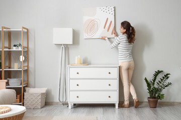 Young woman hanging picture near chest of drawers in stylish room