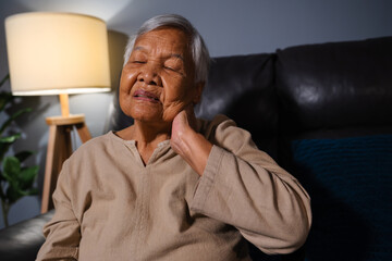 senior woman suffering from neck pain while sitting on sofa in living room at night